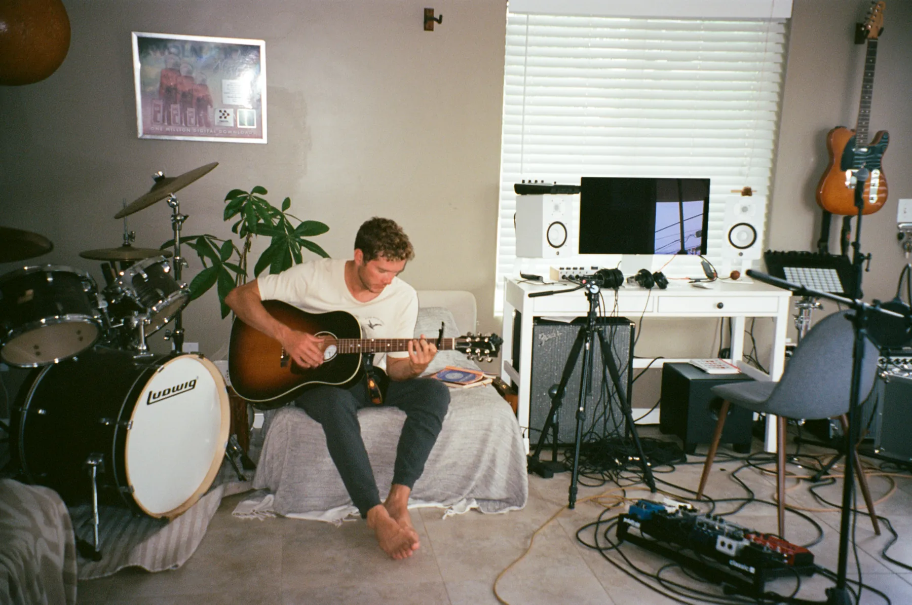Brandon playing acoustic guitar in his home studio in San Diego, surrounded by a drum kit, a Telecaster on the wall, a Fender combo amp, studio monitors and a pedalboard